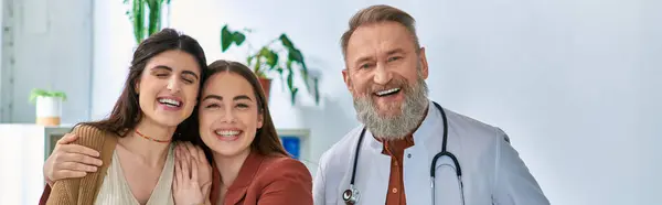 Grey bearded cheerful doctor smiling at camera standing near smiley lgbt couple, ivf concept, banner — Stock Photo
