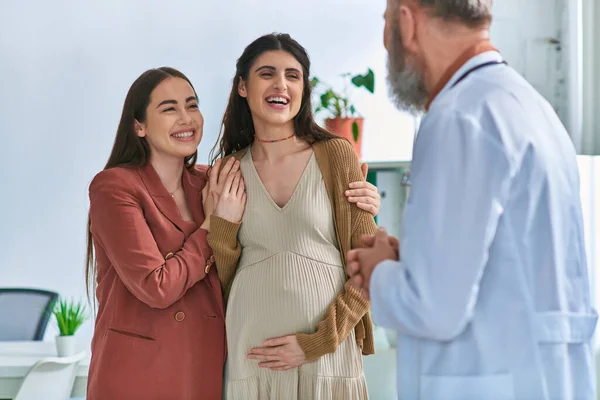Freudiges lesbisches Paar mit Händen auf schwangerem Bauch, das ihren Frauenarzt anlächelt, ivf concept — Stockfoto