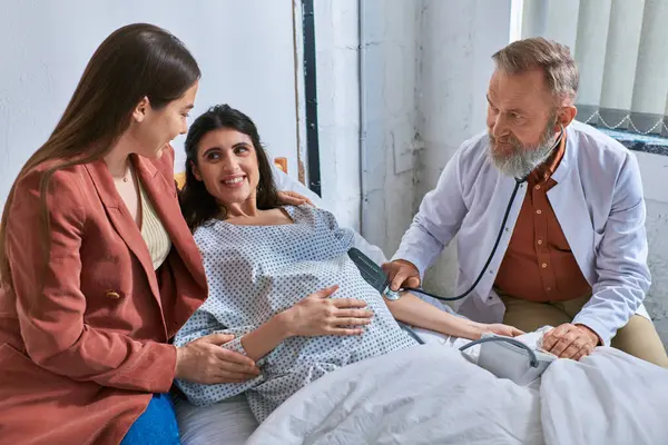 Lgbt casal sorrindo um para o outro enquanto médico medir a pressão na mulher grávida, ivf conceito — Fotografia de Stock