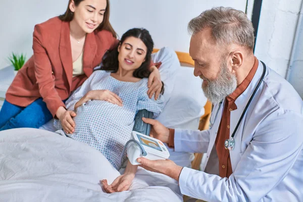 Happy lgbt couple hugging with hand on pregnant belly while doctor measuring pressure, ivf concept — Stock Photo