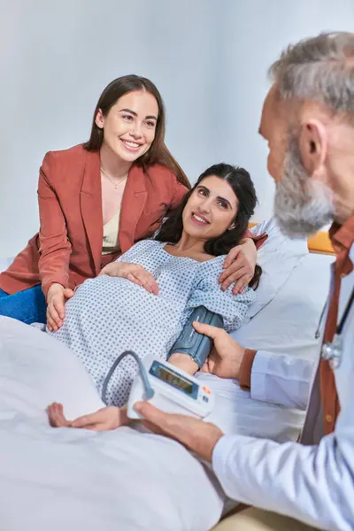 Cheerful lesbian couple hugging and looking at doctor that measuring pressure, ivf concept — Stock Photo