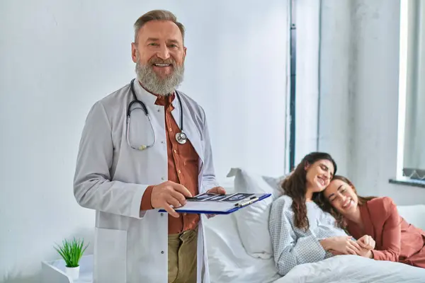 Doctor looking at camera with ultrasound in hands with hugging lgbt couple on backdrop, ivf concept — Stock Photo