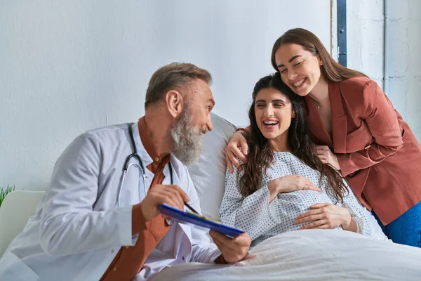 Cheerful doctor looking at happy lesbian couple hugging and smiling sincerely, ivf concept — Stock Photo