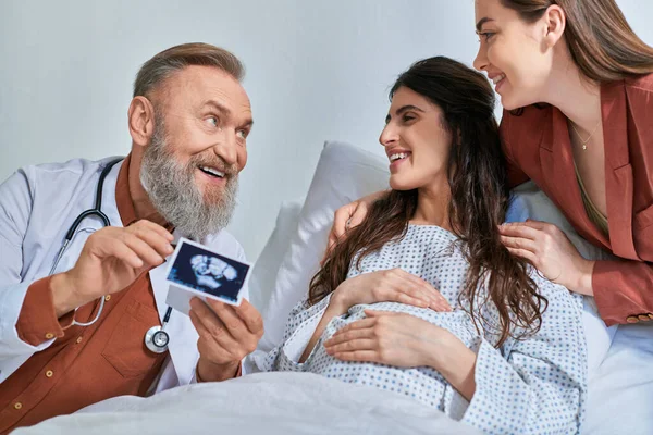 Усміхнений сірий бородатий лікар, що показує ультразвук, щоб обійняти пару lgbt, дивлячись на них, концепція IVf — стокове фото