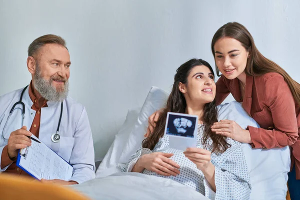 Happy lesbian couple hugging and looking at ultrasound with doctor next to them, ivf concept — Stock Photo