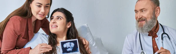 Joyous lesbian couple hugging warmly looking at ultrasound with doctor aside, ivf concept, banner — Stock Photo