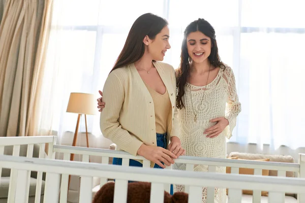 Joyous beautiful lesbian couple standing next to crib with hand on pregnant belly, ivf concept — Stock Photo