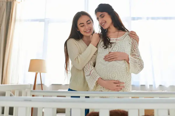 Счастливая лесбийская пара тепло обнимается с рукой на беременном животе, стоя рядом с кроваткой, ivf концепция — стоковое фото