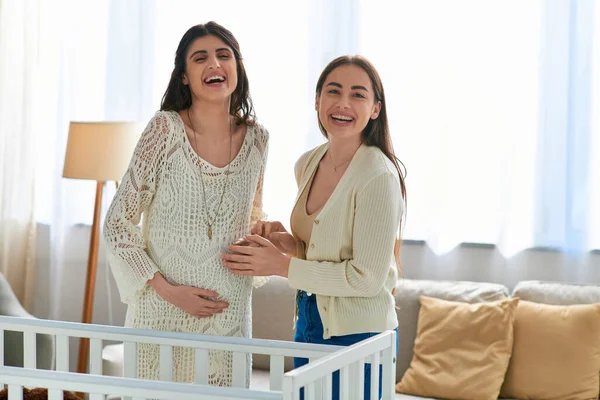 Joyous lgbt couple standing next to crib smiling at camera with hands on pregnant belly, ivf concept — Stock Photo