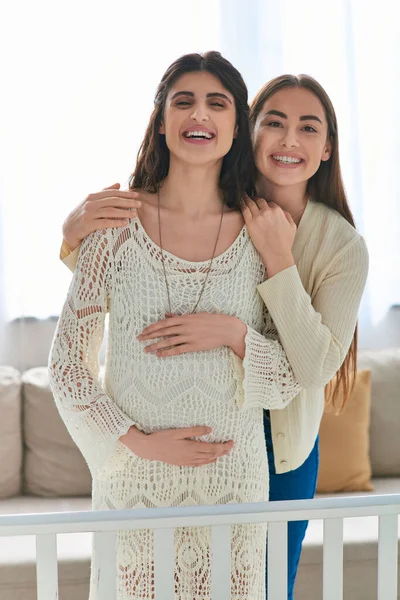 Happy lgbt couple standing next to crib smiling sincerely at camera, hands on belly, ivf concept — Stock Photo