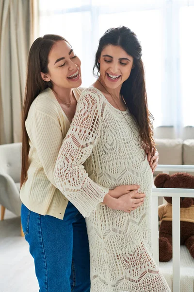 Beautiful lgbt couple hugging warmly with brown teddy bear in crib on backdrop, ivf concept — Stock Photo