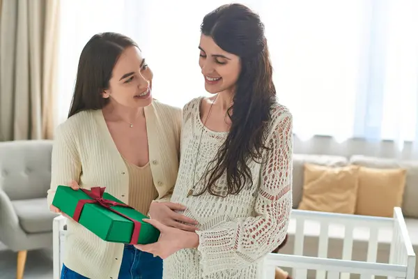 Joyous lesbian couple standing next to crib holding gift with hands on pregnant belly, ivf concept — Stock Photo