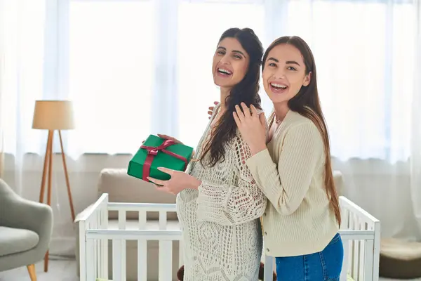 Cheerful lesbian couple standing next to crib with gift in hands and smiling at camera, ivf concept — Stock Photo