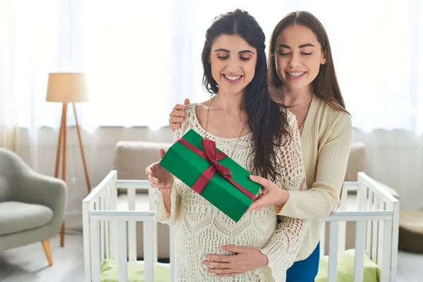 Pretty lesbian couple standing next to crib holding present with hand on pregnant belly, ivf concept — Stock Photo