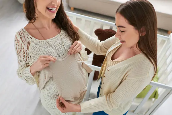 Cropped view of expecting lesbian couple with singlet in hands with crib on backdrop, ivf concept — Stock Photo