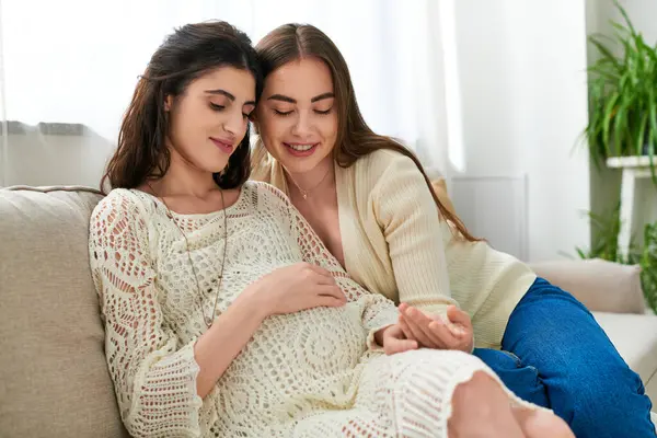 Hermosa embarazada pareja lesbiana abrazándose en sofá con la mano en el vientre embarazada, concepto ivf - foto de stock