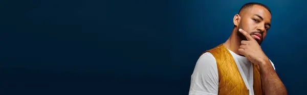 Young man in stylish yellow vest posing on dark blue backdrop, hand to face, fashion concept, banner — Stock Photo