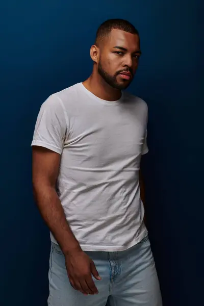 Handsome young man in white t-shirt and jeans posing on blue backdrop looking away, fashion concept — Stock Photo
