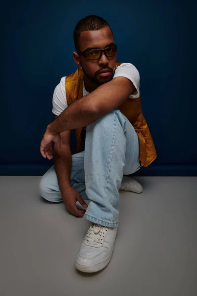 Афроамериканський чоловік присідає і позує на темно-синьому фоні з головою на руці, концепція моди — стокове фото