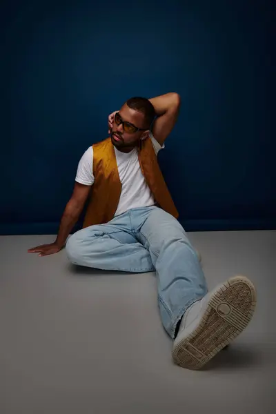 Красивый мужчина в стильном жилете и солнцезащитных очках, сидящих на полу, рука об шею, концепция моды — стоковое фото