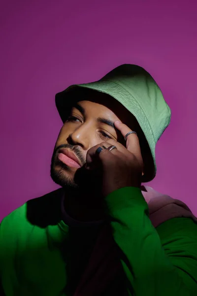 Homme réfléchi en panama vert avec des anneaux touchant son front sur fond violet, concept de mode — Photo de stock