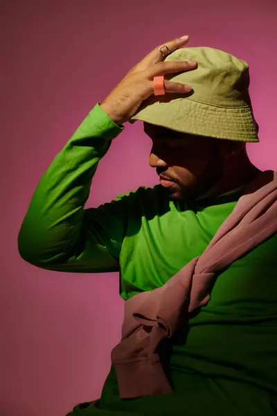 Красивый бородатый мужчина с кольцами позирует в профиль с рукой на зеленой панаме, концепция моды — стоковое фото