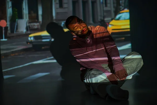 Jeune homme afro-américain incliné légèrement sur le sol avec les mains sur les jambes croisées, concept de mode — Photo de stock