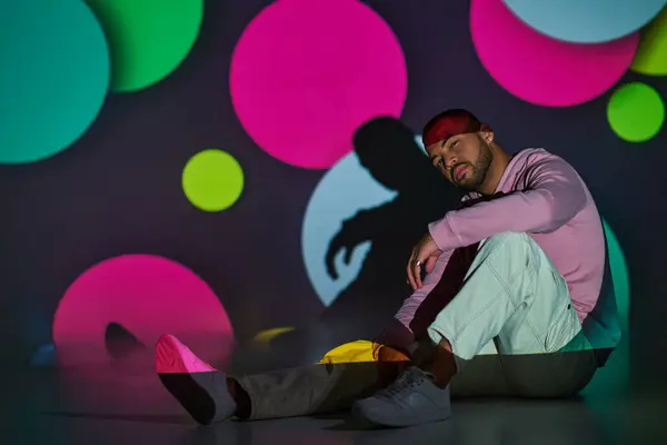 Schöner junger Mann sitzt leicht lächelnd auf dem Boden im digitalen Projektorlicht, Modekonzept — Stockfoto