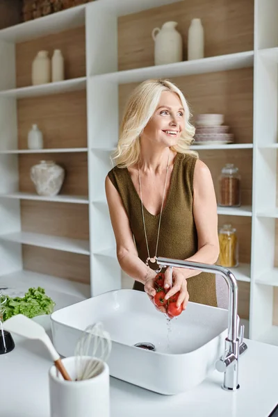 Веселая женщина средних лет моет свежие и спелые помидоры черри и улыбается на современной кухне — стоковое фото
