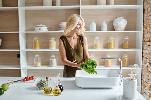 Mulher alegre de meia-idade com cabelo loiro lavar alface fresca na pia da cozinha, cozinhar em casa — Fotografia de Stock