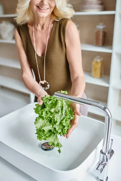 Vista recortada de mujer alegre de mediana edad con cabello rubio lavando lechuga fresca en fregadero de cocina - foto de stock
