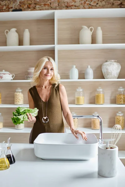 Привлекательная вегетарианка средних лет с светлыми волосами моющая свежий салат в кухонной раковине — стоковое фото