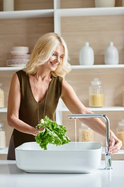 Mujer vegetariana positiva de mediana edad con cabello rubio lavando lechuga fresca, tiro vertical - foto de stock