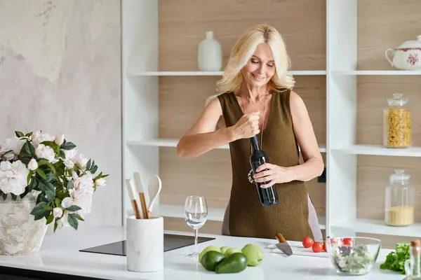 Joyeuse femme d'âge moyen avec des cheveux blonds ouverture bouteille de vin près de légumes frais sur le comptoir — Photo de stock
