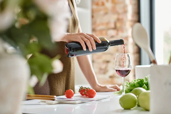 Femme cultivée verser du vin rouge dans un verre près de pommes fraîches et tomates cerises sur le comptoir — Photo de stock