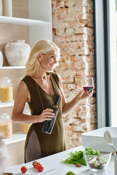 Mulher de meia idade despreocupada com o cabelo loiro segurando garrafa e vidro com vinho tinto na cozinha — Fotografia de Stock