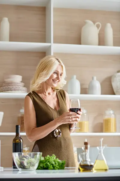Красивая женщина средних лет с светлыми волосами держа стакан с красным вином и улыбаясь на современной кухне — стоковое фото