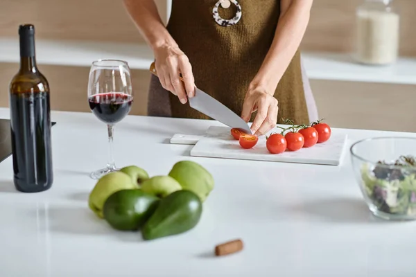 Mujer recortada cortando tomates cherry frescos cerca de un vaso de vino tinto, aguacate y manzanas verdes - foto de stock