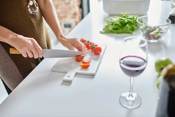 Tiro parcial de mulher cortando tomates cereja perto de vidro de vinho tinto e alface em tigela transparente — Fotografia de Stock