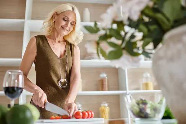 Feliz rubia de mediana edad mujer cortando tomates cherry cerca de un vaso de vino tinto en la cocina moderna - foto de stock