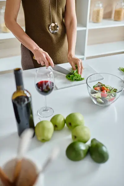 Mujer recortada cortando lechuga fresca y haciendo ensalada de verduras cerca de un vaso de vino tinto en la encimera - foto de stock