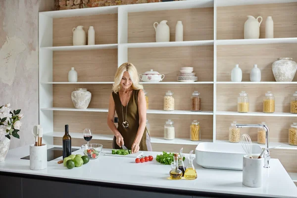 Modern kitchen, cheerful middle aged woman with blonde hair making salad from fresh ingredients — Stock Photo