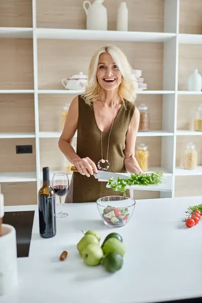 Збуджена жінка середнього віку робить салат зі свіжих інгредієнтів на сучасній кухні, домашня кухня — стокове фото
