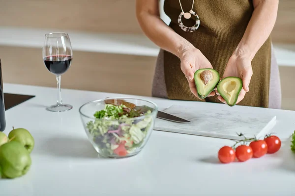 Donna ritagliata in possesso di avocado fresco metà vicino insalata in ciotola e bicchiere di vino rosso sul piano di lavoro — Foto stock