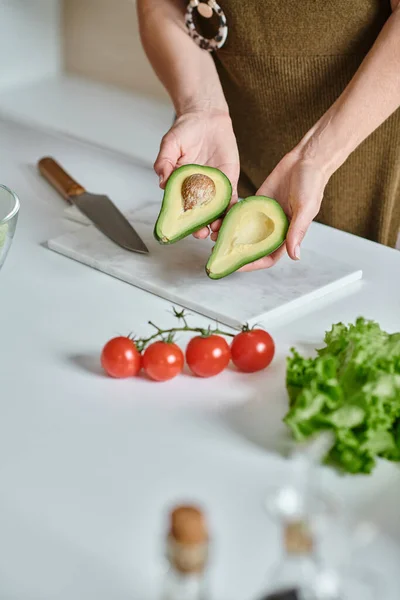 Cropped woman holding fresh avocado halves near cherry tomatoes, lettuce and knife on countertop — Stock Photo