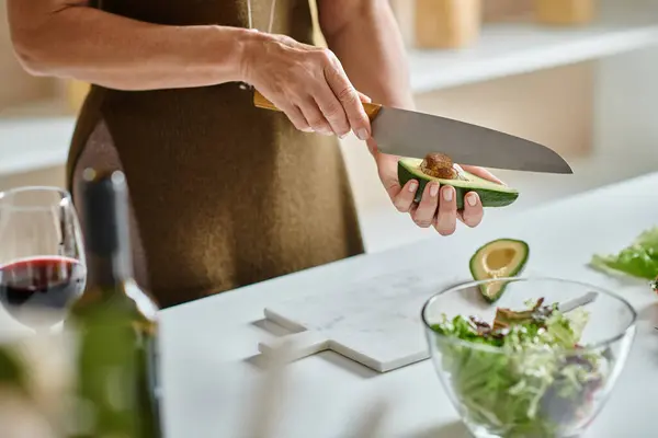 Частичный выстрел, женщина режет свежий авокадо рядом с помидорами черри и салатом в миске, домашняя кухня — стоковое фото