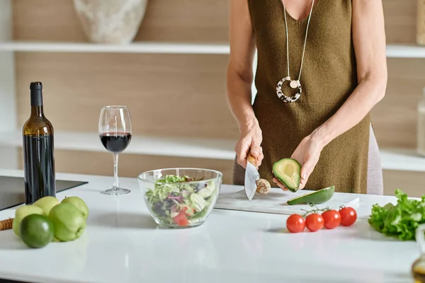 Partial shot, woman cutting ripe avocado near fresh ingredients and red wine, home cooking — Stock Photo