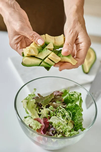 Женщина держит нарезанный спелый авокадо возле салата в стеклянной чаше, крупным планом женские руки — стоковое фото