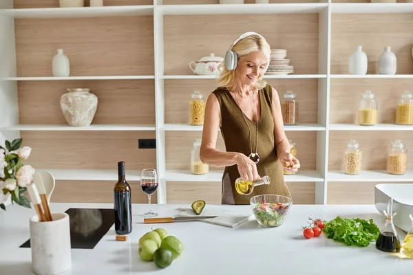 Joyeuse femme d'âge moyen dans des écouteurs sans fil faire de la salade près de la bouteille et un verre de vin rouge — Photo de stock