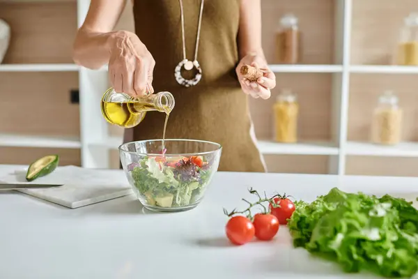 Tiro recortado de la mujer que vierte el aceite de oliva en un recipiente de vidrio con ensalada cerca de las verduras en la encimera - foto de stock